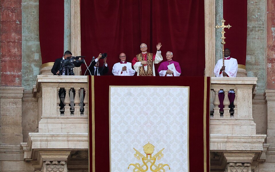 El papa León XIV saluda desde el balcón central de la basílica de San Pedro, acompañado de varios clérigos.
