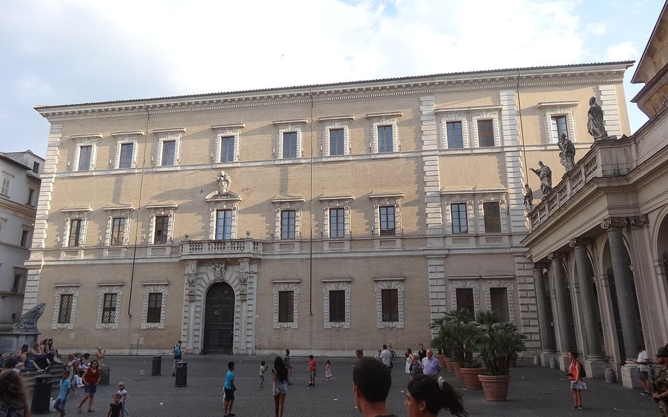 Fachada de un palacio romano de color crema con muchas ventanas, frente a una plaza con gente caminando.